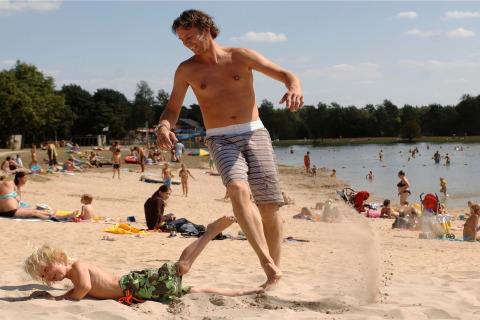 Ein Mann und ein Kind spielen lachend am Sandstrand eines Sees, umgeben von Menschen am Wasser.