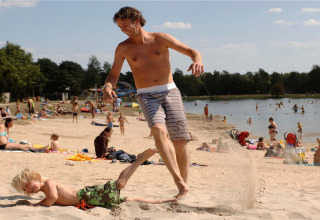 Un homme et un enfant jouent et rient sur une plage de sable au bord d’un lac, entourés de vacanciers.