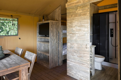Interno di una tenda safari con mobili in legno, letti a castello, TV, tavolo da pranzo e bagno privato.