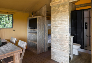 Intérieur d’une tente safari avec meubles en bois, lits superposés, TV, table à manger et salle de bain.