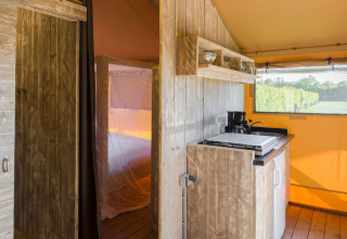 Interno di una tenda safari con angolo cottura, pareti in legno e vista su siepi verdi all'esterno.