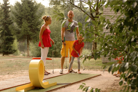 En familie spiller minigolf udendørs i en grøn park, omgivet af træer og afslappet naturstemning.