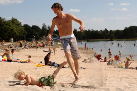 Mand sparker sand ved strand med barn, mennesker bader og slapper af ved søen i Forest Park 't Wolfsven 13.
