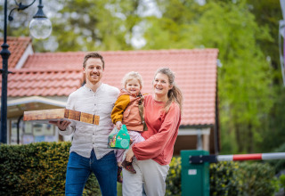 Famille souriante devant une tiny house au toit rouge au Holiday Park De Rimboe & De Woeste Hoogte, Pays-Bas.