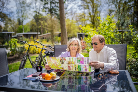 Älteres Paar sitzt an einem Tisch im Freien, studiert eine Karte bei Glamping-Unterkünften, Fahrräder im Hintergrund.