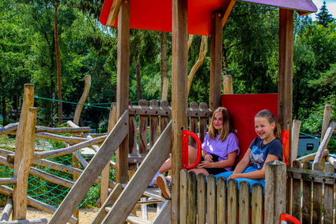 Twee meisjes ontspannen en lachen in een houten speelhuisje op de speeltuin van een glampingverblijf.