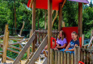 Zwei Mädchen sitzen und lachen in einem Holzspielhaus auf einem Glamping-Spielplatz im Grünen.