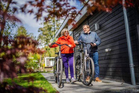 Pareja mayor con bicicletas sonríe frente a su alojamiento glamping rodeado de naturaleza verde.