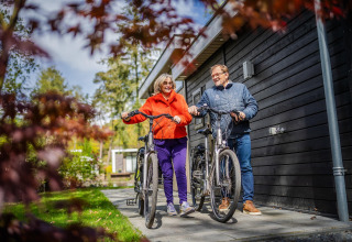 Senior couple with bicycles smiling in front of their modern glamping accommodation, enjoying nature.