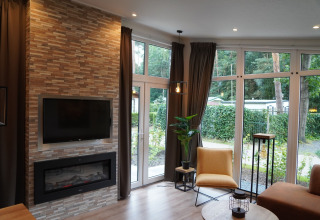 Modernes Wohnzimmer in einer Lodge mit Kamin, TV, großen Fenstern und Gartenblick in den Niederlanden.