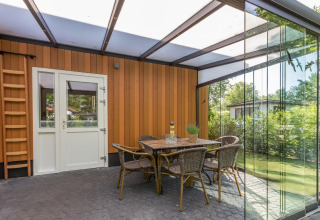 Overdekt terras met glazen wanden, eettafel en rotanstoelen bij Residence De Eese 3 lodge, Nederland.