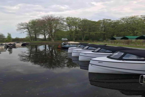 Både fortøjet ved en søbred i Rekreatiepark Idskenhuizen 11 med træer og hytter i baggrunden, Holland.