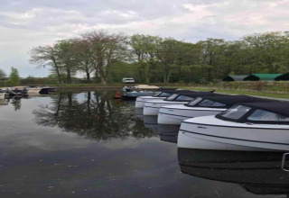 Både fortøjet ved en søbred i Rekreatiepark Idskenhuizen 11 med træer og hytter i baggrunden, Holland.