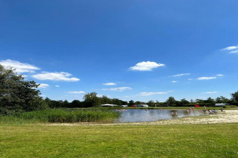 Udsigt over en sø med græsplæne og folk, blå himmel og hytter i baggrunden i Recreation Park Idskenhuizen.