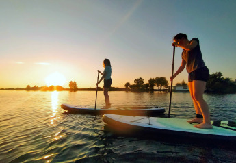 To personer står på paddleboards ved solnedgang ved Chalet «Imke» i Holland, omgivet af vand og træer.