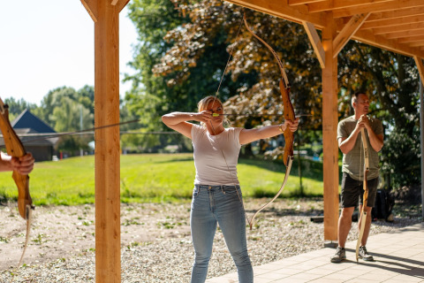 Kvinde skyder med bue og pil under overdækning på en glamping-plads, mens andre deltager i baggrunden.