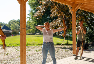 Kvinde skyder med bue og pil under overdækning på en glamping-plads, mens andre deltager i baggrunden.