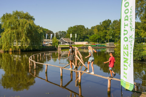 Børn krydser en smal træbro over en dam ved Hunzepark 13 lodge i Holland på en solrig dag.