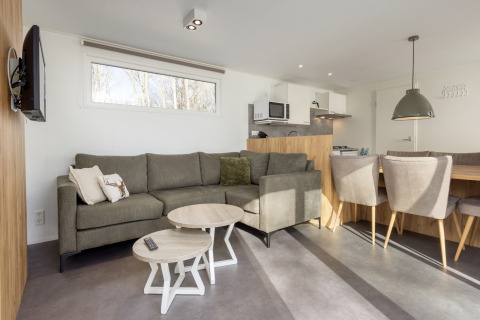 Moderne lodge Hunzepark 13 i Holland med grå sofa, spiseplads, åbent køkken og store vinduer.