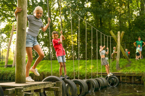 Børn leger på en dækbro ved Hunzepark 13 i Nederlandene, omgivet af grøn natur og klar himmel.