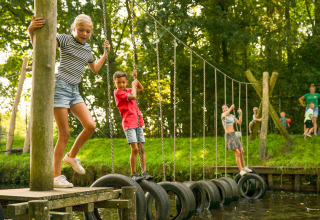 Børn leger på en dækbro ved Hunzepark 13 i Nederlandene, omgivet af grøn natur og klar himmel.