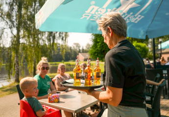 Serveerster brengt bier naar een gezin aan een buitentafel onder een parasol bij een glamping-accommodatie.