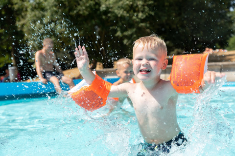 Et smilende barn med orange svømmevinger leger i poolen ved Hunzepark 13-lodgen i Nederlandene.