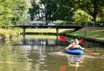 Twee mensen kajakken op een rustige gracht bij Hunzepark 15 in Nederland, met groene bomen en een brug.