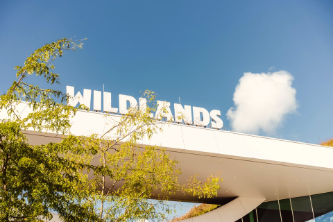 Entrance of WILDLANDS Adventure Zoo Emmen with large white sign and tree branches under blue sky.