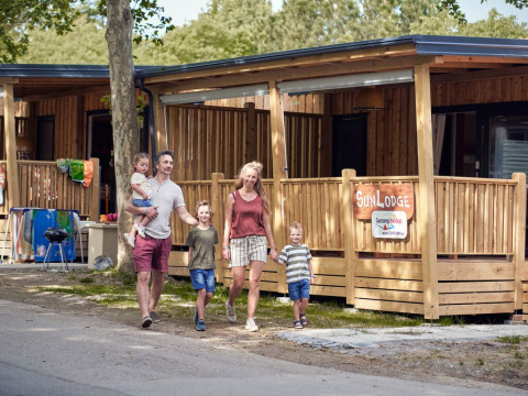 Familia caminando frente a SunLodge Mobile Home Katsura en el camping Pra'delle Torri en Italia, cabañas de madera atrás.