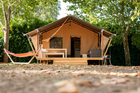 The Nomad safari tent with wooden deck, hammock, and outdoor furniture beneath lush green trees outdoors.
