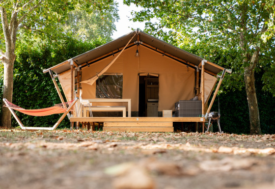 The Nomad safari tent with wooden deck, hammock, and outdoor furniture beneath lush green trees outdoors.