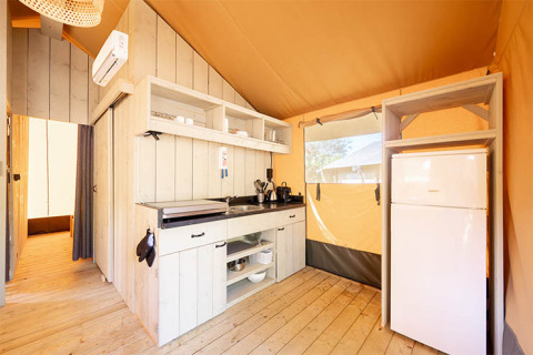 Intérieur d’une tente safari avec petite cuisine, sol en bois, réfrigérateur blanc et étagères ouvertes.