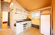 Interieur van een safaritent met compacte keuken, houten vloer, witte koelkast en open rekken.