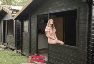 Woman sits in the window of a tiny black wooden house at Wecamp Cudillero in Spain, with red table and chairs outside.