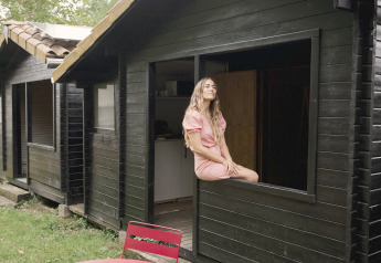 Woman sits in the window of a tiny black wooden house at Wecamp Cudillero in Spain, with red table and chairs outside.