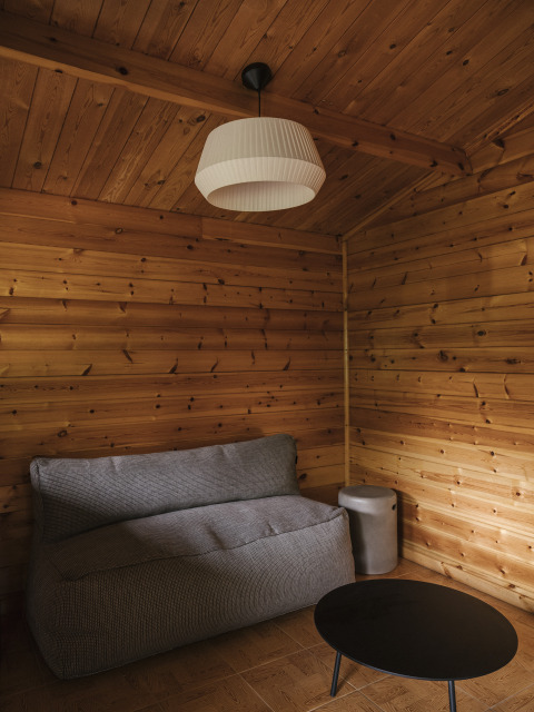 Cabaña de glamping con paredes de madera, sofá gris, lámpara moderna y mesa redonda negra pequeña.