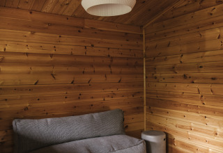 Cabaña de glamping con paredes de madera, sofá gris, lámpara moderna y mesa redonda negra pequeña.