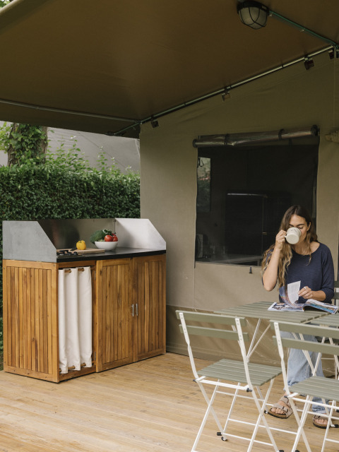 Une femme boit du café et lit à une table sous une tente safari avec cuisine extérieure à Wecamp Cudillero.