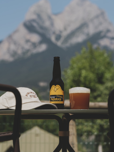 Een fles bier, glas en pet op een tafel met vervaagde bergen op de achtergrond bij Wecamp Pedraforca, Catalonië.