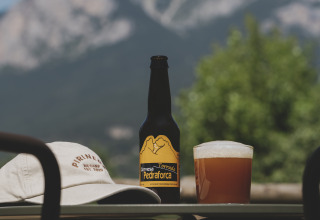 Een fles bier, glas en pet op een tafel met vervaagde bergen op de achtergrond bij Wecamp Pedraforca, Catalonië.