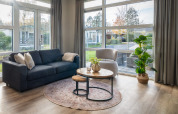 Moderne opholdsstue i Forest Lodge med store vinduer, sofa, lænestol og udsigt til haven i Nederlandene.