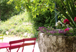 Colorful chairs and a table beside a stone planter with flowers in a lush camping area surrounded by greenery.