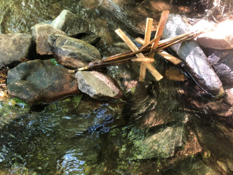A handmade water wheel built from sticks and rocks rests in a clear stream at Moulin de Chaules campsite, France.