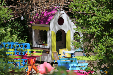 Area giochi all'aperto per bambini con casetta, tavolini, sedie e giochi da giardino immersa nel verde.
