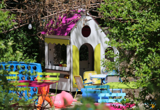 Area giochi all'aperto per bambini con casetta, tavolini, sedie e giochi da giardino immersa nel verde.