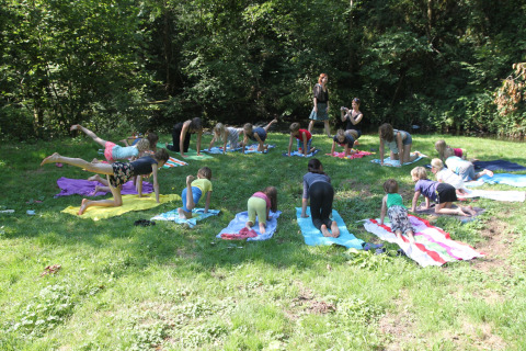Children practice outdoor yoga on colorful towels at a green campsite in a nature park in France.