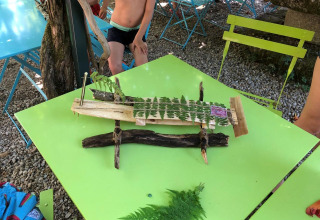 Children playing outdoors at a table with a handmade boat of wood and leaves at a campsite in France.