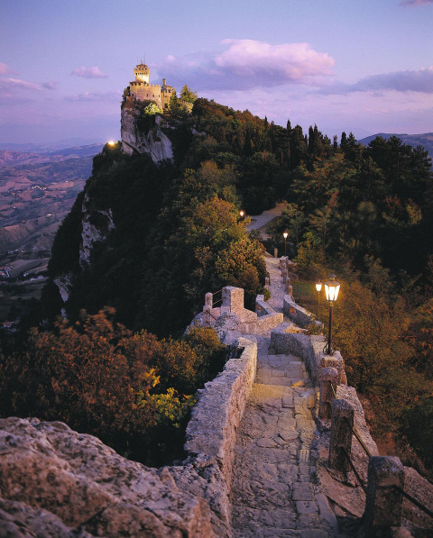 Chemin de pierre éclairé menant à un château perché à San Marino, entouré de forêts et vues au crépuscule.