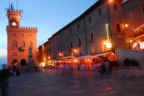 Aftenbillede af en oplyst plads med historiske bygninger og restauranter i Repubblica di San Marino.